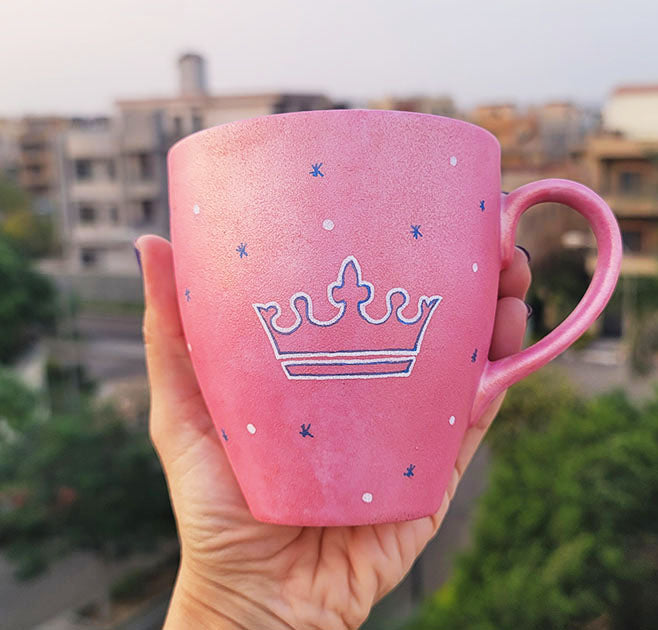 A Hand - painted shimmery pink mug with A Crown painted in blue