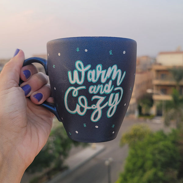 A Shimmery blue hand painted mug with the words "Warm and Cozy " in silver