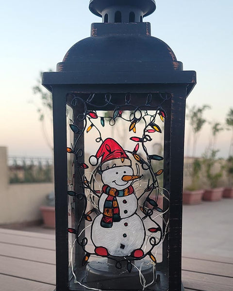 A Black Vintage Lantern with a snowman painted on the glass and festive lights above him