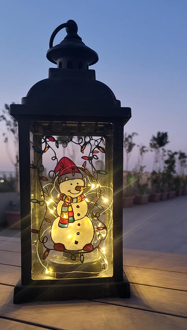 A Black Vintage Lantern with a snowman painted on the glass and festive lights above him, shown at night when the lantern lights are on