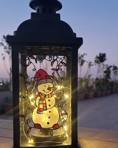 A Black Vintage Lantern with a snowman painted on the glass and festive lights above him, shown at night when the lantern lights are on