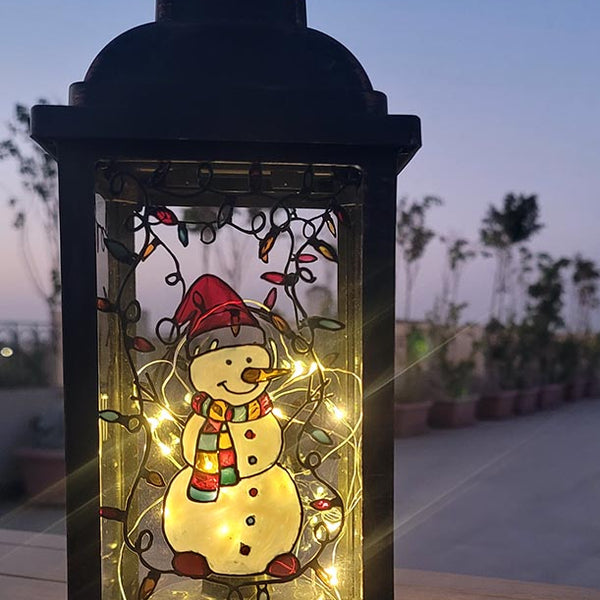 A Black Vintage Lantern with a snowman painted on the glass and festive lights above him, shown at night when the lantern lights are on