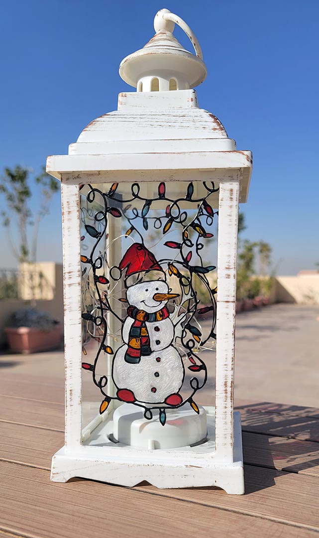 A White Vintage Lantern with a snowman painted on the glass and festive lights above him