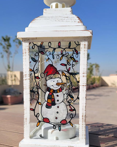 A White Vintage Lantern with a snowman painted on the glass and festive lights above him