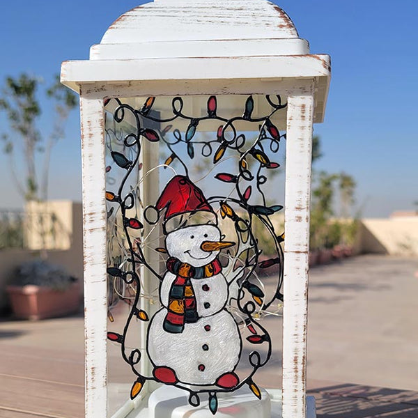 A White Vintage Lantern with a snowman painted on the glass and festive lights above him