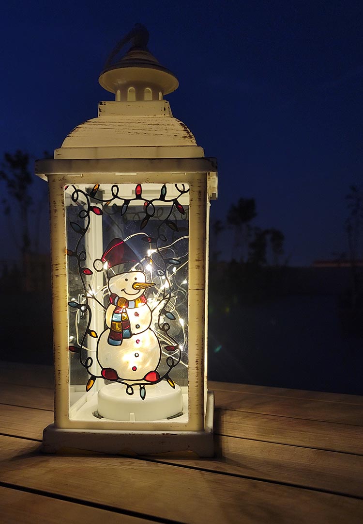 A White Vintage Lantern with a snowman painted on the glass and festive lights above him, shown at night when the lantern lights are on