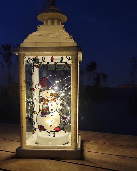 A White Vintage Lantern with a snowman painted on the glass and festive lights above him, shown at night when the lantern lights are on