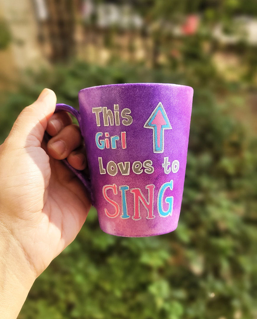 A Purple Hand painted mug with the sentence "This Girl loves to sing" and an arrow pointed up. All written in white and colored in with grey blue and pink.