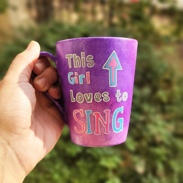 A Purple Hand painted mug with the sentence "This Girl loves to sing" and an arrow pointed up. All written in white and colored in with grey blue and pink.