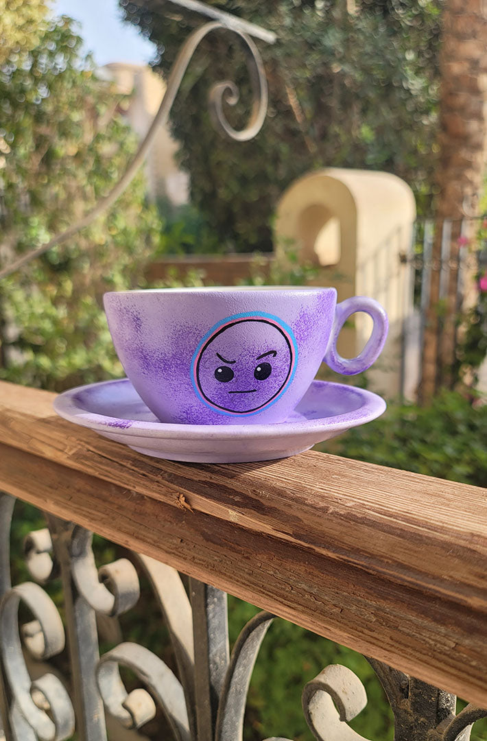 A Lilac hand painted cup and saucer with the cup having the raised eyebrow emoji in black and outlined with pink and blue