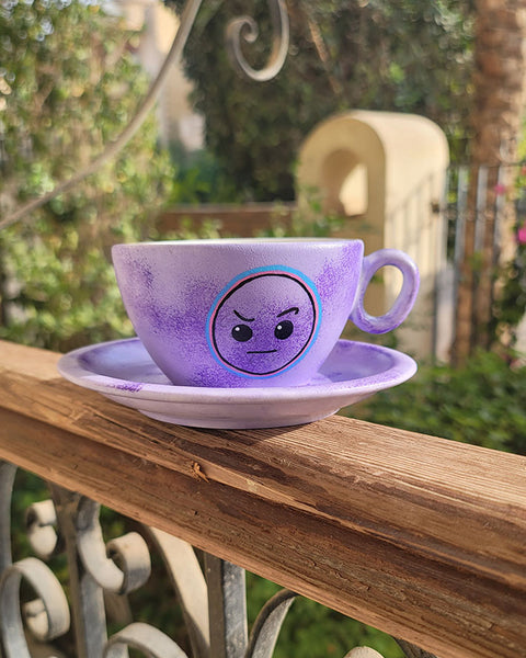 A Lilac hand painted cup and saucer with the cup having the raised eyebrow emoji in black and outlined with pink and blue