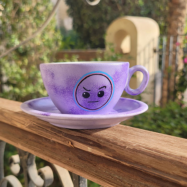 A Lilac hand painted cup and saucer with the cup having the raised eyebrow emoji in black and outlined with pink and blue