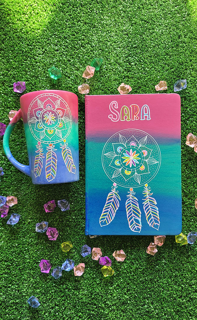 A gradient hand painted mug and notebook (pink, green, blue) with a dreamcatcher in white and colored in pink, purple, orange, yellow, green and blue and the name sara