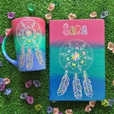 A gradient hand painted mug and notebook (pink, green, blue) with a dreamcatcher in white and colored in pink, purple, orange, yellow, green and blue and the name sara