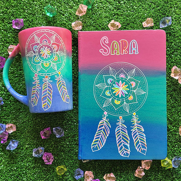 A gradient hand painted mug and notebook (pink, green, blue) with a dreamcatcher in white and colored in pink, purple, orange, yellow, green and blue and the name sara