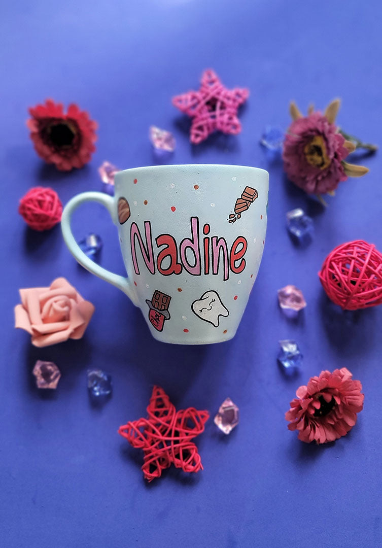 A Baby Blue hand painted mug with the name nadine and chocolate bars, tooth, toothbrush and chocolate pieces scattered all over.