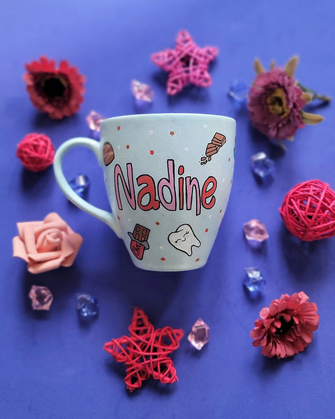 A Baby Blue hand painted mug with the name nadine and chocolate bars, tooth, toothbrush and chocolate pieces scattered all over.