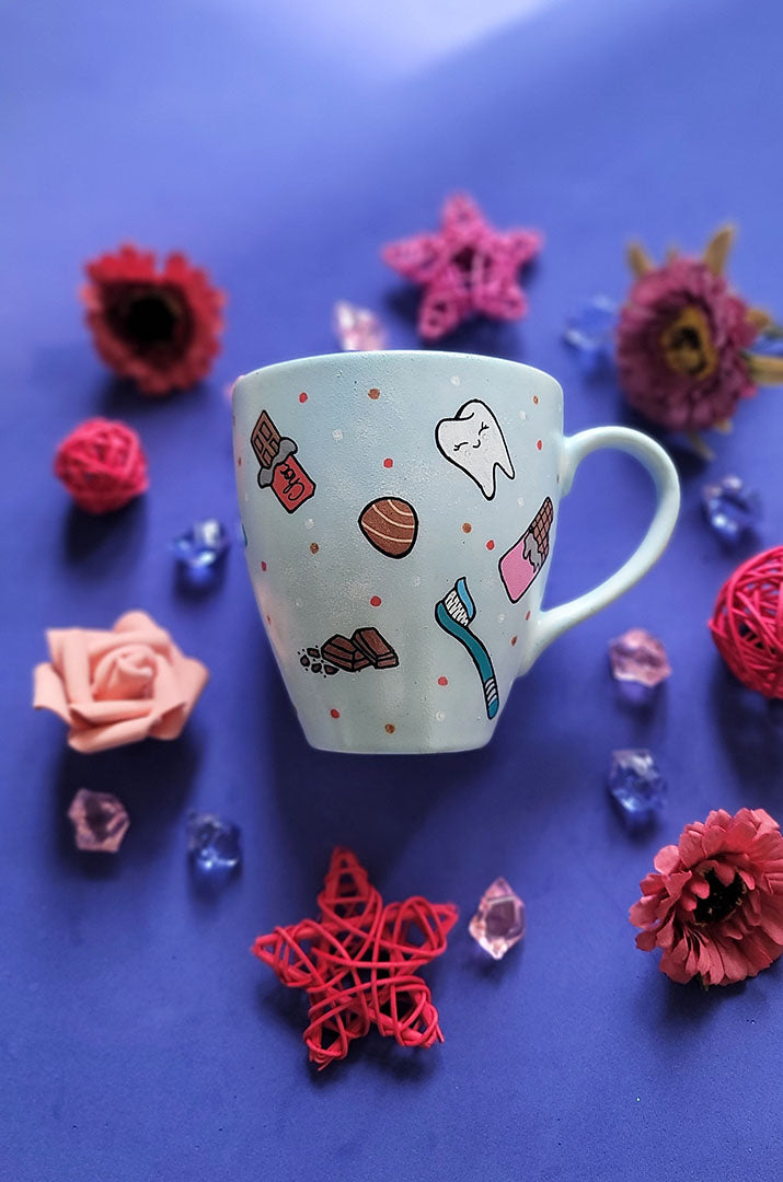 A Baby Blue hand painted mug with chocolate bars, tooth, toothbrush and chocolate pieces scattered all over.