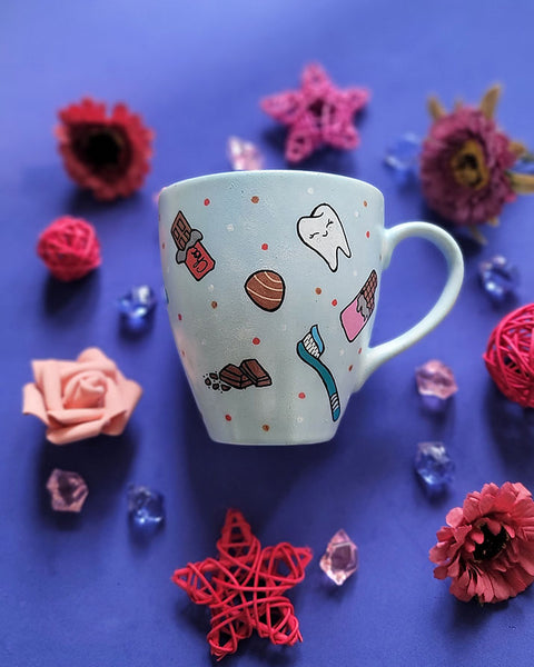 A Baby Blue hand painted mug with chocolate bars, tooth, toothbrush and chocolate pieces scattered all over.