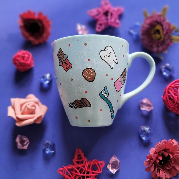 A Baby Blue hand painted mug with chocolate bars, tooth, toothbrush and chocolate pieces scattered all over.