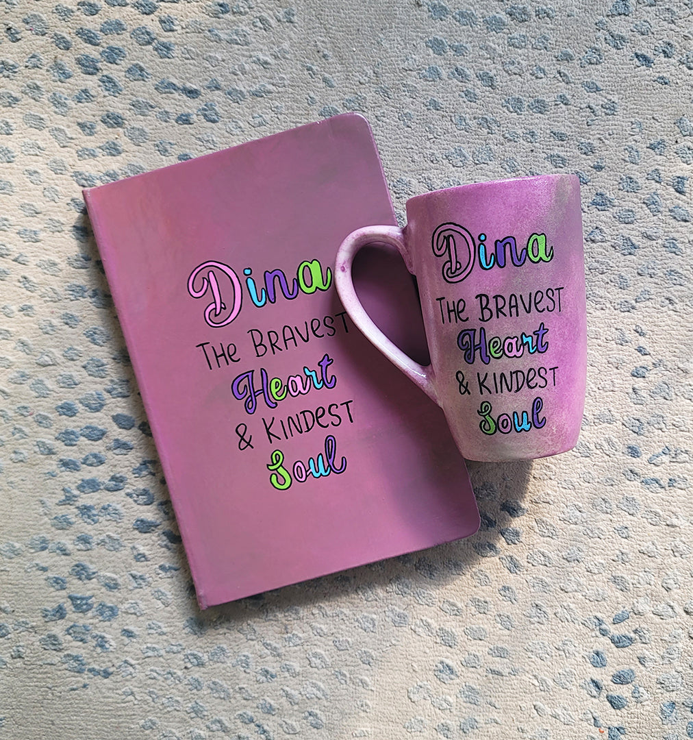 A Pastel Pink Hand painted gift set of mug and notebook with the name Dina wirtten on top and under it the sentence "the bravest heart and kindest Soul" all wirtten in black and colored in purple, pink, green and blue.