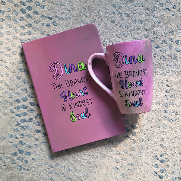 A Pastel Pink Hand painted gift set of mug and notebook with the name Dina wirtten on top and under it the sentence "the bravest heart and kindest Soul" all wirtten in black and colored in purple, pink, green and blue.