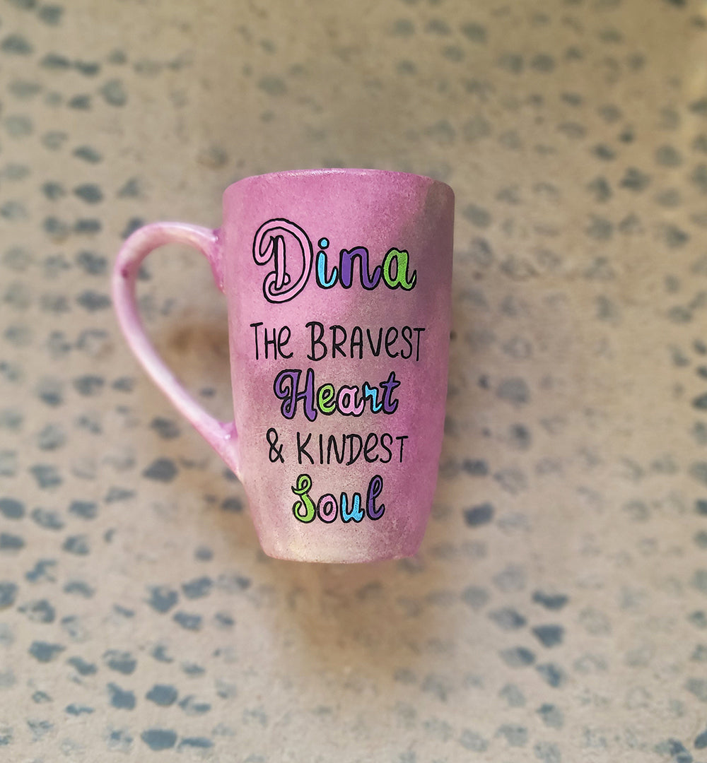 A Pastel Pink Hand painted mug with the name Dina wirtten on top and under it the sentence "the bravest heart and kindest Soul" all wirtten in black and colored in purple, pink, green and blue.