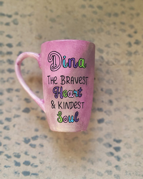 A Pastel Pink Hand painted mug with the name Dina wirtten on top and under it the sentence "the bravest heart and kindest Soul" all wirtten in black and colored in purple, pink, green and blue.