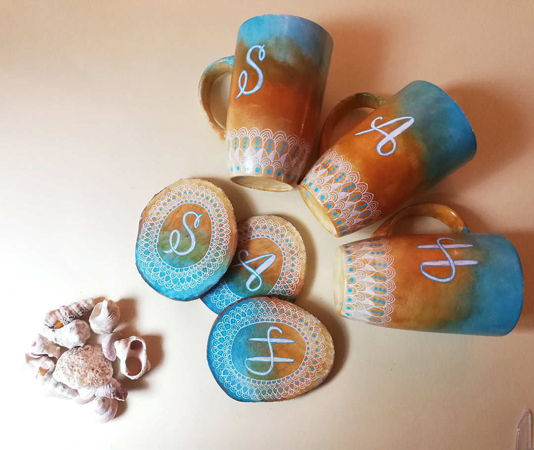 A Picture showing three hand painted mugs and three matching coasters are colored in gradient of "blue-bej-white" with patterns drawn all around the bottom in white and each mug and coaster has a letter written on it "S","A" and "H"