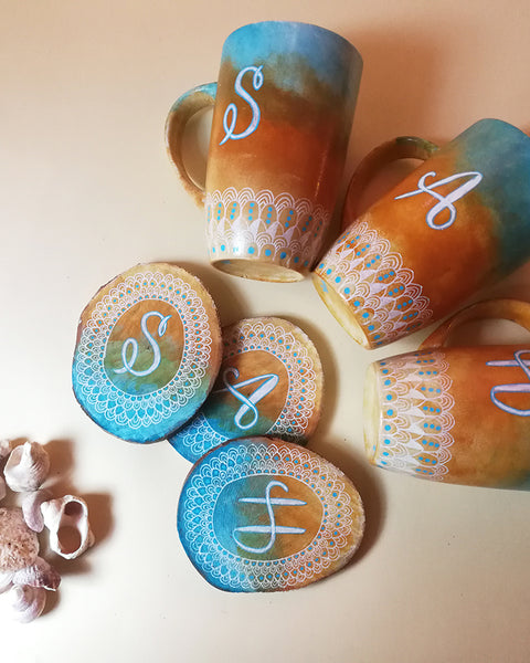 A Picture showing three hand painted mugs and three matching coasters are colored in gradient of "blue-bej-white" with patterns drawn all around the bottom in white and each mug and coaster has a letter written on it "S","A" and "H"