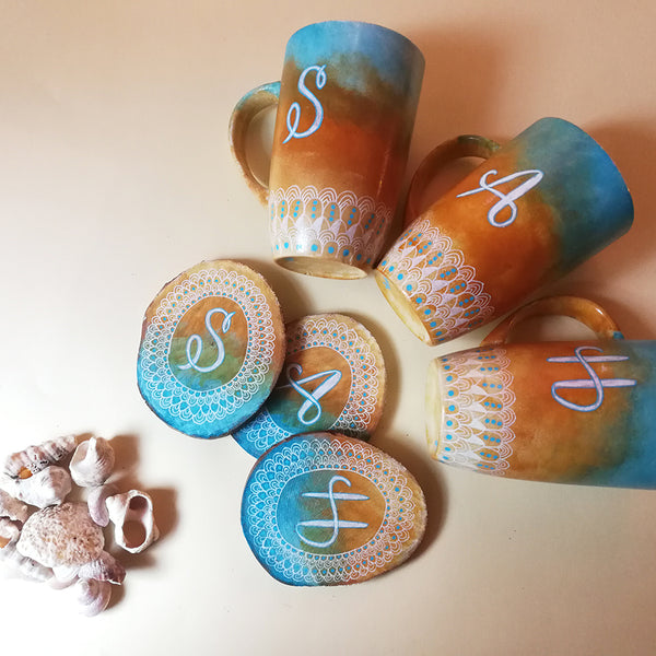 A Picture showing three hand painted mugs and three matching coasters are colored in gradient of "blue-bej-white" with patterns drawn all around the bottom in white and each mug and coaster has a letter written on it "S","A" and "H"