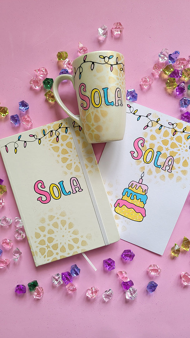 A Gift Set of A matching mug, notebook and card . All handpainted in ivory with geometric patterns in gold, and have festive lights all over the top. The notebook and mug have the name "Sola" and the card has the name sola and a cake design on it