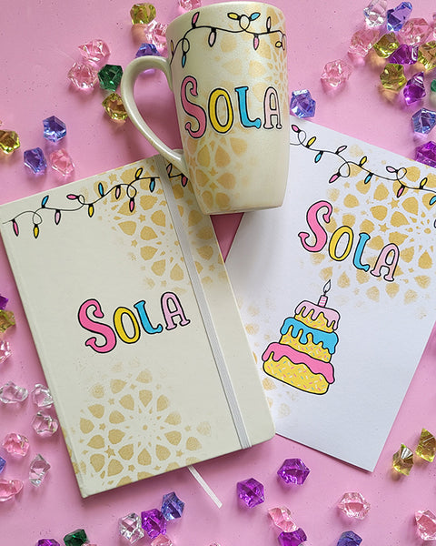 A Gift Set of A matching mug, notebook and card . All handpainted in ivory with geometric patterns in gold, and have festive lights all over the top. The notebook and mug have the name "Sola" and the card has the name sola and a cake design on it