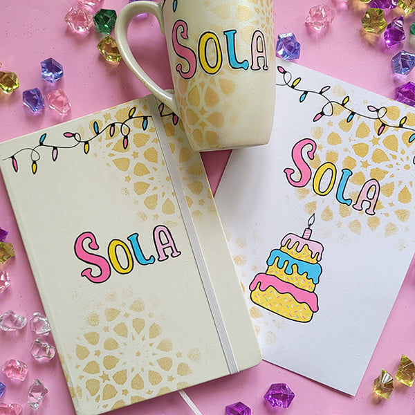 A Gift Set of A matching mug, notebook and card . All handpainted in ivory with geometric patterns in gold, and have festive lights all over the top. The notebook and mug have the name "Sola" and the card has the name sola and a cake design on it