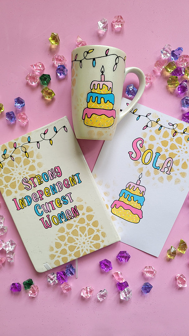 A Gift Set of A matching mug, notebook and card . All handpainted in ivory with geometric patterns in gold, and have festive lights all over the top. The notebook has the words "Strong Independent Cutest Woman" the mug has a cake design and the card has the name sola and a cake design on it