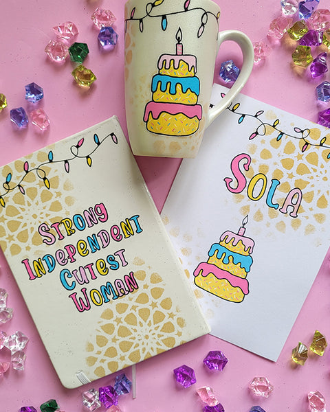 A Gift Set of A matching mug, notebook and card . All handpainted in ivory with geometric patterns in gold, and have festive lights all over the top. The notebook has the words "Strong Independent Cutest Woman" the mug has a cake design and the card has the name sola and a cake design on it