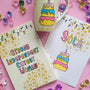 A Gift Set of A matching mug, notebook and card . All handpainted in ivory with geometric patterns in gold, and have festive lights all over the top. The notebook has the words "Strong Independent Cutest Woman" the mug has a cake design and the card has the name sola and a cake design on it