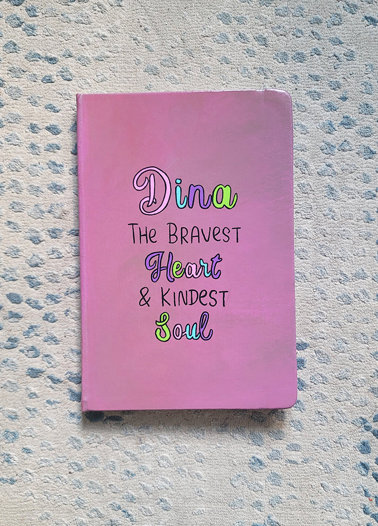 A Pastel Pink Hand painted Notebook with the name Dina wirtten on top and under it the sentence "the bravest heart and kindest Soul" all wirtten in black and colored in purple, pink, green and blue.