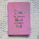 A Pastel Pink Hand painted Notebook with the name Dina wirtten on top and under it the sentence 