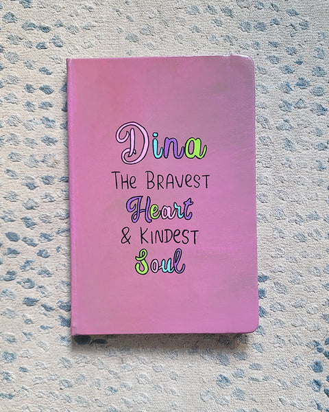 A Pastel Pink Hand painted Notebook with the name Dina wirtten on top and under it the sentence "the bravest heart and kindest Soul" all wirtten in black and colored in purple, pink, green and blue.