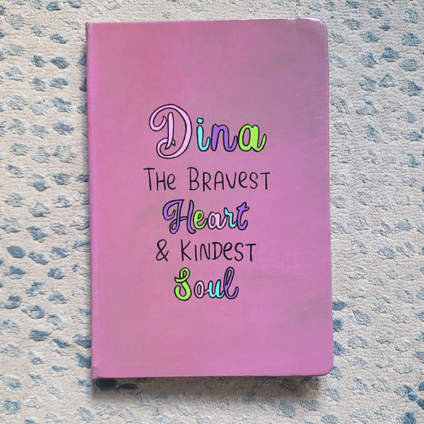 A Pastel Pink Hand painted Notebook with the name Dina wirtten on top and under it the sentence "the bravest heart and kindest Soul" all wirtten in black and colored in purple, pink, green and blue.