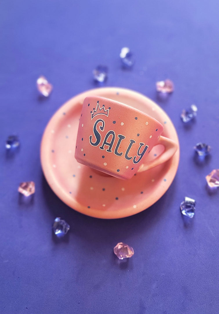 A Hand - painted shimmery pink cup and saucer with the name Sally with a crown in blue and white and blue dots scattered all over