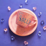 A Hand - painted shimmery pink cup and saucer with the name Sally with a crown in blue and white and blue dots scattered all over