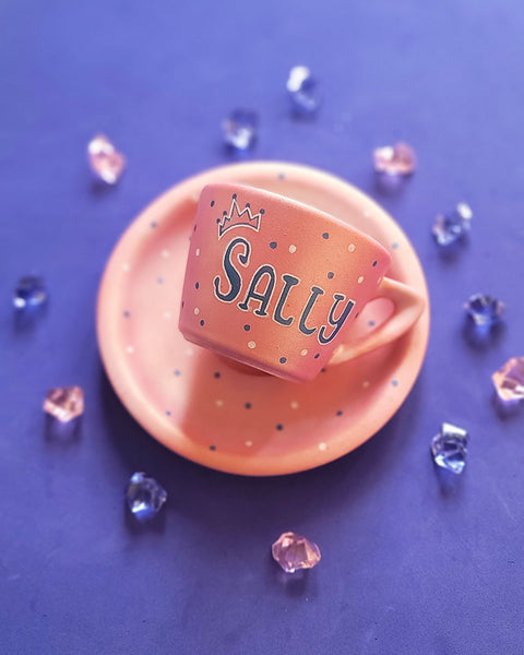 A Hand - painted shimmery pink cup and saucer with the name Sally with a crown in blue and white and blue dots scattered all over