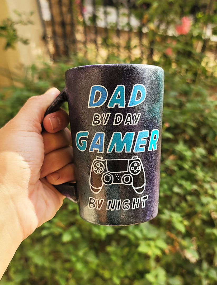 A Black Sky Hand painted mug with the sentence " Dad by day, Gamer by night" and a joystick drawn under the word gamer.