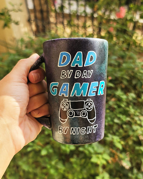 A Black Sky Hand painted mug with the sentence " Dad by day, Gamer by night" and a joystick drawn under the word gamer.