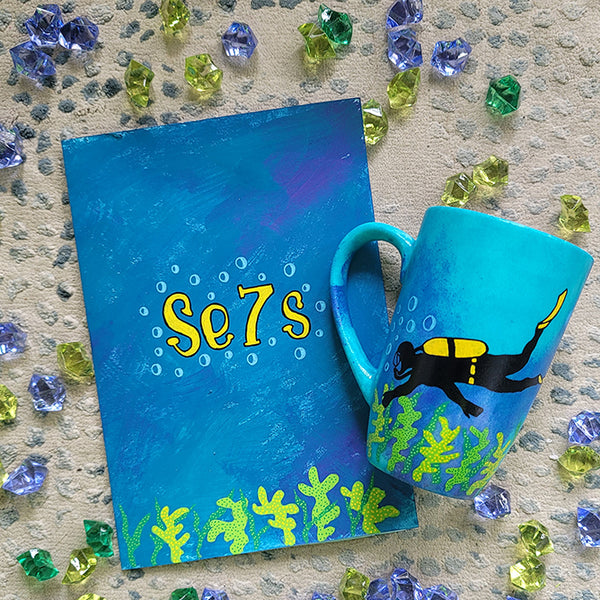 A Gift Set of a mug and matching card all in blue galaxy color and with coral reefs on the bottom. the card has the name Se7s and the mug has a diver 