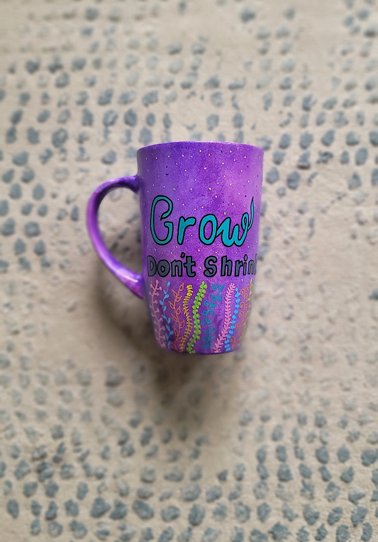 A Purple hand painted mug with the words "grow, Don't Shrink" and plants rising from the bottom of the mug to the third of height all around