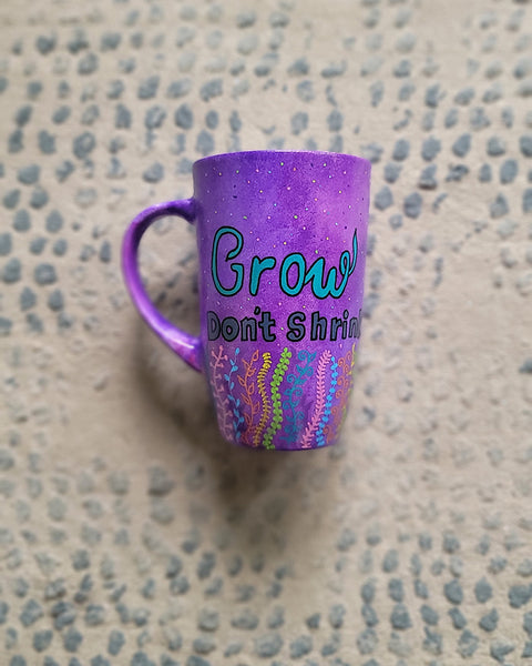 A Purple hand painted mug with the words "grow, Don't Shrink" and plants rising from the bottom of the mug to the third of height all around