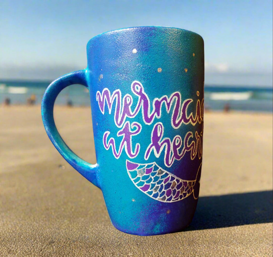 A blue hand painted mug with the words "Mermaid at heart" with a mermaid tail painted below it